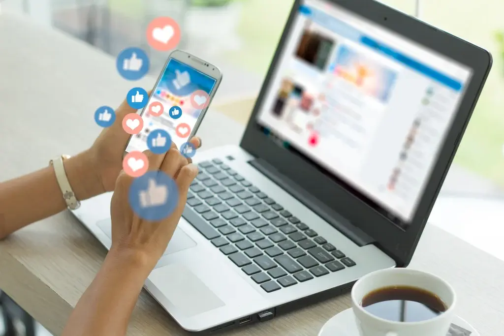 A person using a smartphone and laptop simultaneously, with social media reaction icons (likes and hearts) floating from the phone screen. The image highlights the social media engagement risks that regulated businesses face, such as compliance challenges and uncontrolled content exposure.
